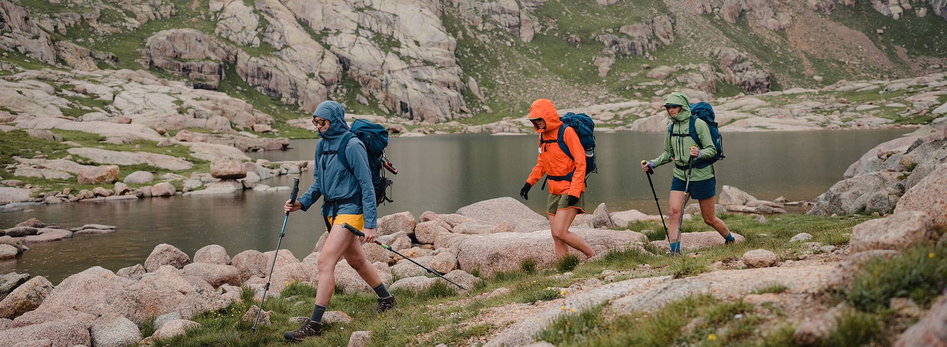 troje ludzi w kurtkach przeciwdeszczowych idzie brzegiem górskiego jeziora