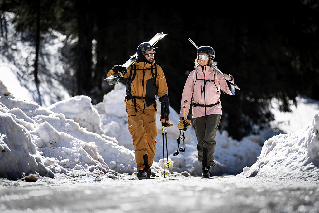kobieta i mężczyzna idą drogą niosą na ramionach narty w ręku kije narciarskie mają na sobie spodnie salewa 